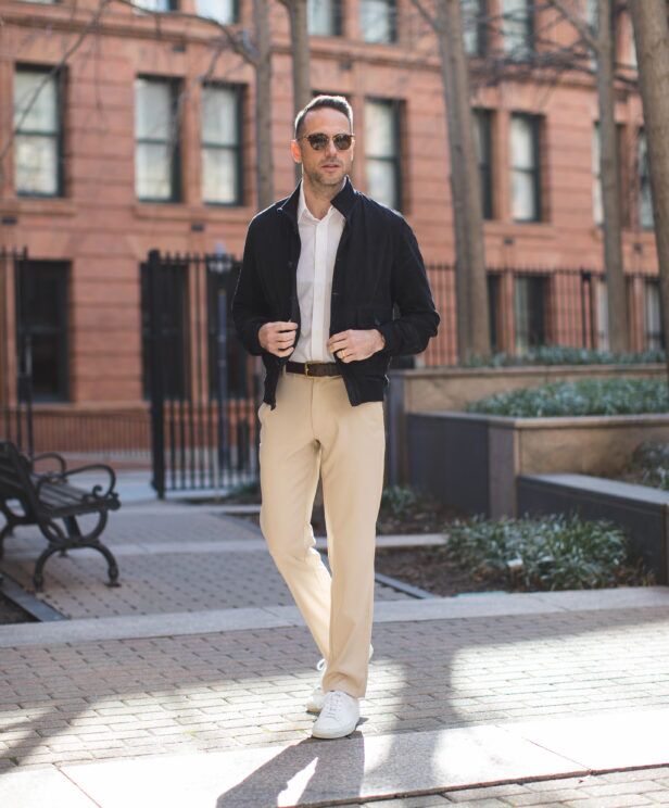 Navy suede blouson jacket with white shirt, cream trousers and white sneakers, worn by a model on a city street