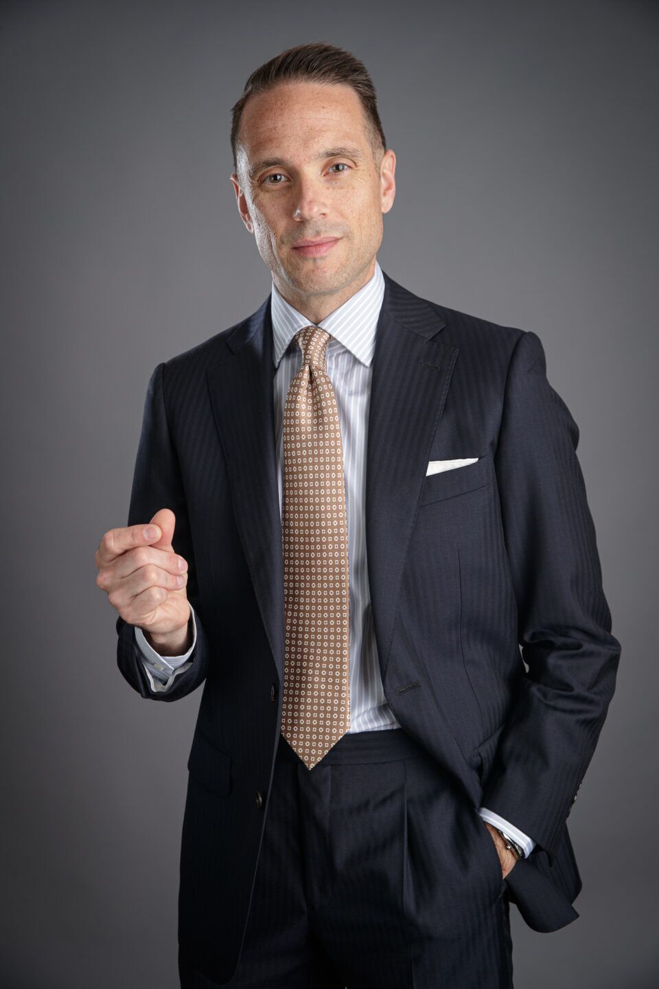 Navy herringbone wool suit with grey striped shirt, brown medallion silk tie and white pocket square, worn by a model
