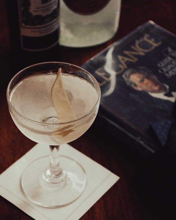 Coupe glass martini with lemon twist on a bar napkin beside a book and bottles, a styled lifestyle scene