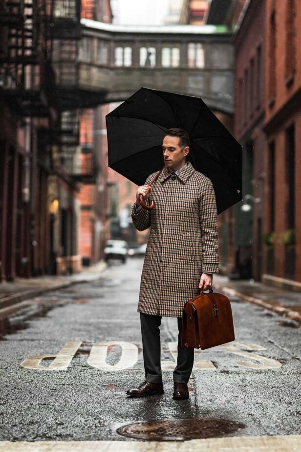 Brown houndstooth wool overcoat with grey trousers, brown briefcase and black umbrella, worn by a model on a rainy street