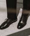 Black leather cap toe oxfords with black trousers, worn by a model on concrete