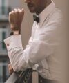 White formal dress shirt with french cuffs, gold knot cufflinks and black bow tie, worn by a model on a balcony