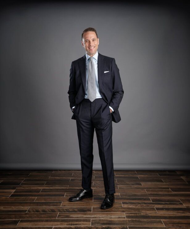 Dark navy striped two-piece suit with light blue shirt, grey check tie and white pocket square, worn by a model