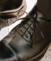 Black leather cap toe oxfords with straight bar lacing, worn with dark trousers, a close-up detail
