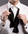 Black silk self-tie butterfly bowtie untied over a white pleated formal shirt with onyx studs and cufflinks, worn by a model