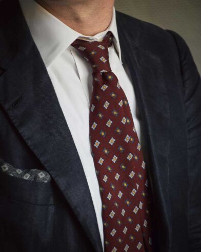 Burgundy silk necktie with blue and gold medallions, worn with a white shirt and navy sport coat, close-up on a model