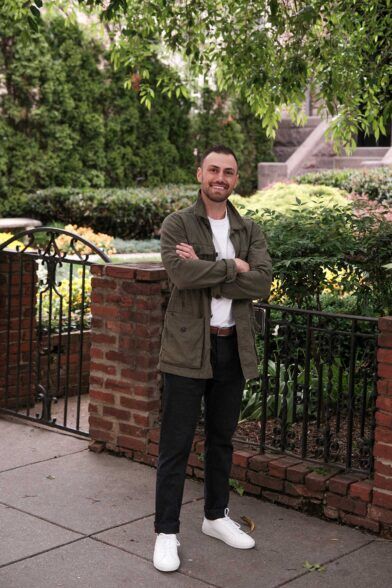 Olive cotton field jacket with white tee, black chinos and white sneakers, worn by a model in a garden setting