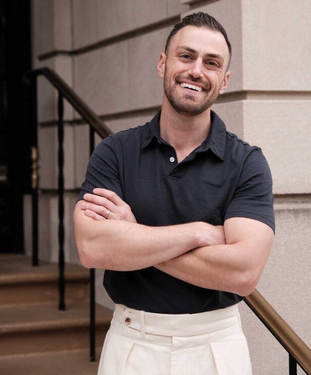 Navy sueded jersey short-sleeve polo with cream pleated trousers, worn by a model on stone steps