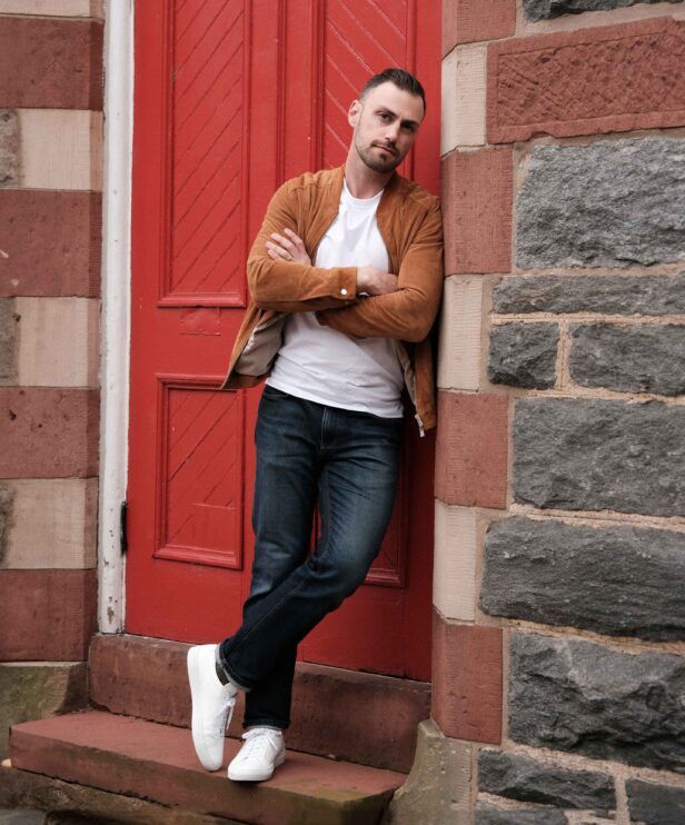 Tan suede bomber jacket with white t-shirt, dark jeans and white sneakers, worn by a model against a red door
