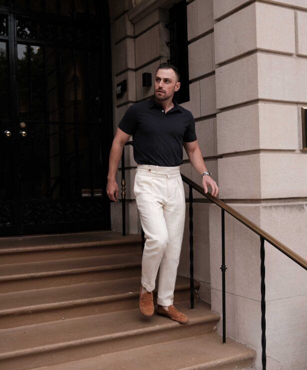 Black sueded jersey polo with cream pleated linen trousers and tan suede loafers, worn by a model on steps