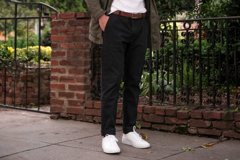 Black cuffed chinos with brown leather belt, olive field jacket and white sneakers, worn by a model on a sidewalk