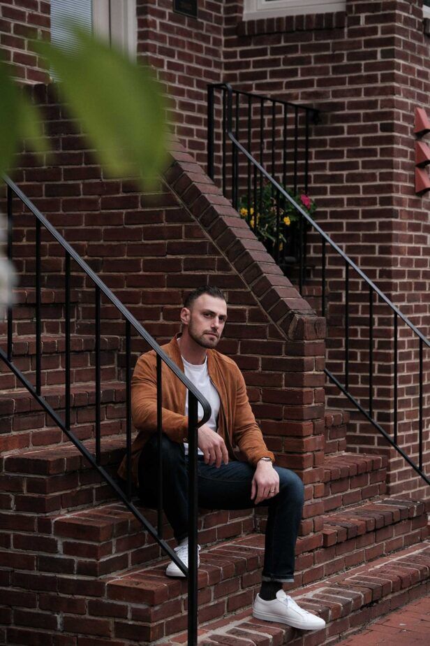 Dark indigo jeans with tan suede blouson, white tee and white sneakers, worn by a model on brick steps