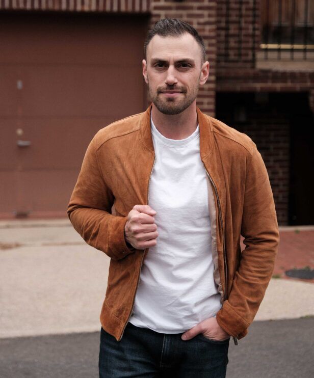 White crewneck tee with tan suede zip bomber jacket and dark denim jeans, worn by a model on a city street