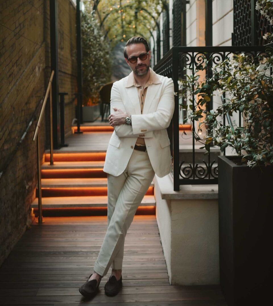 Cream linen suit with camel knit polo and brown loafers, worn by a model on a lit garden staircase
