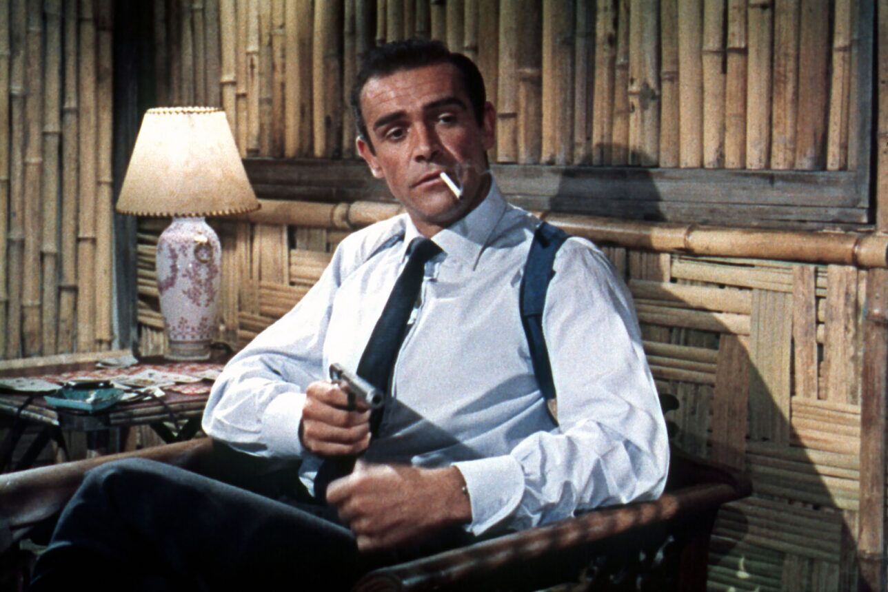 Pale blue dress shirt with dark navy tie, shoulder holster and dark trousers, worn by a seated man in a film still