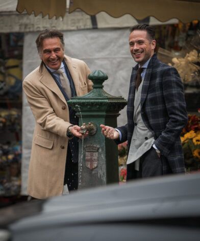 Camel wool overcoat and navy checked topcoat with grey waistcoat, at a Milan street fountain