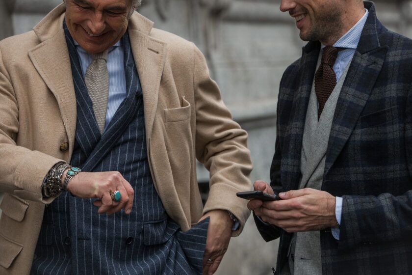 Camel wool overcoat over a navy chalk stripe double-breasted suit beside a navy plaid coat, on a street