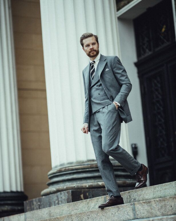Grey flannel three-piece suit with double-breasted waistcoat, striped tie and brown oxfords, worn by a model on steps