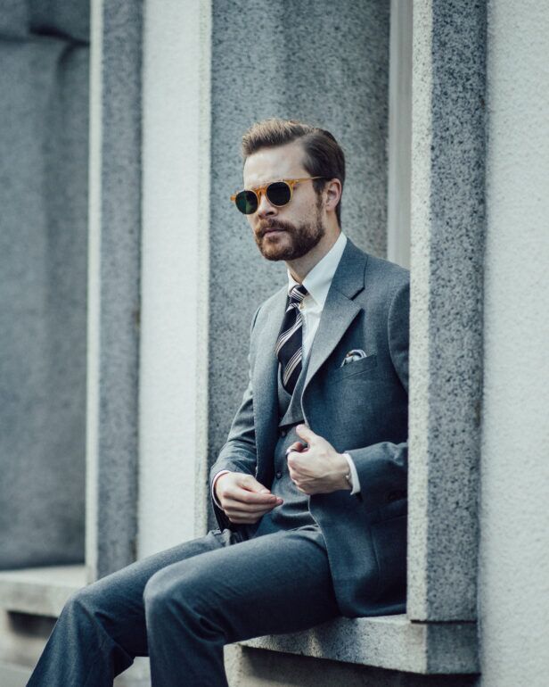 Grey flannel three-piece suit with white shirt, navy striped tie and pocket square, worn by a model seated outdoors