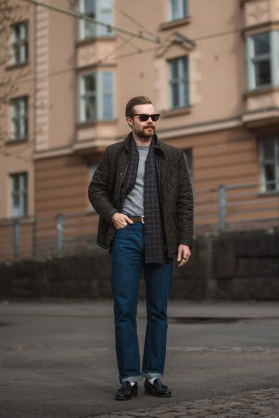 Olive quilted field jacket with grey sweater, patterned silk scarf and blue jeans, worn by a model on a city street
