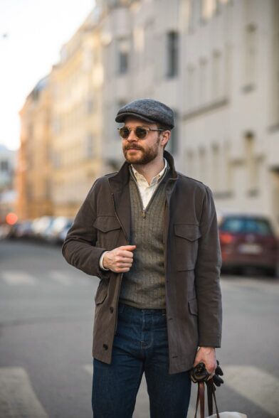 Brown suede field jacket with olive cable-knit half-zip sweater and a tweed flat cap, on a city street