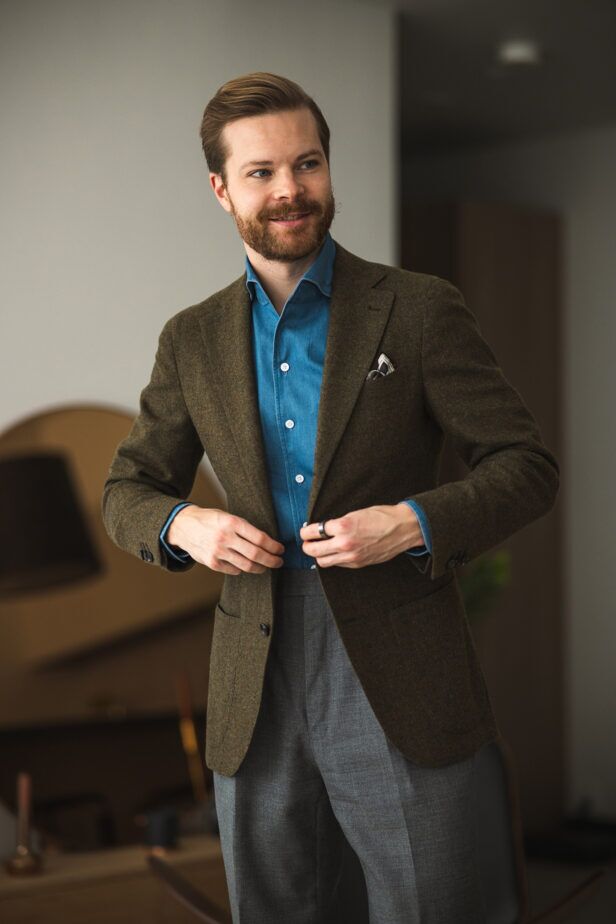 Olive green wool sport coat with blue denim shirt, grey flannel trousers and white pocket square, worn by a model