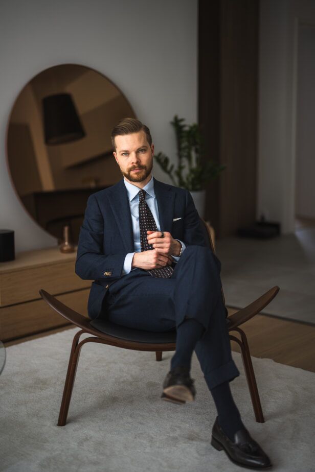Navy flannel suit with blue striped shirt, brown medallion silk tie and black loafers, worn by a seated model