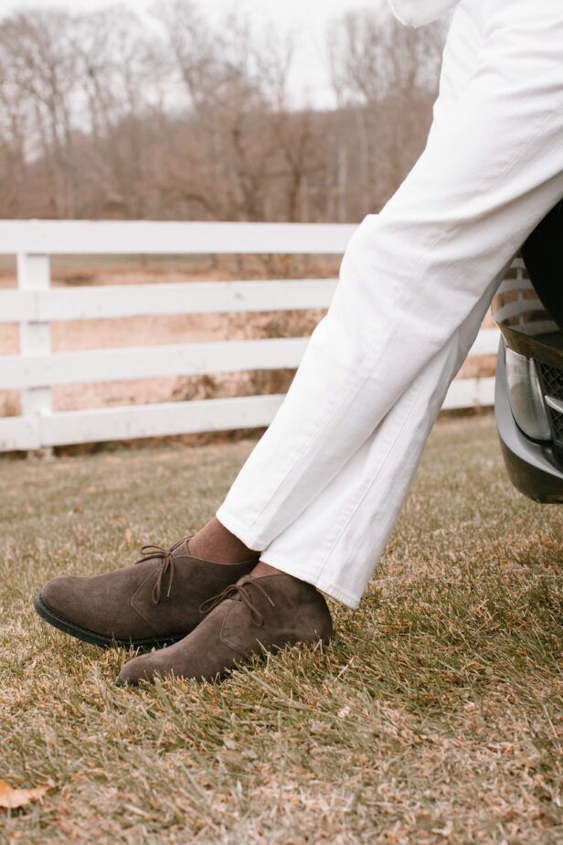 Brown suede chukka boots with white jeans and tan socks, worn by a model on grass, a styled outdoor scene