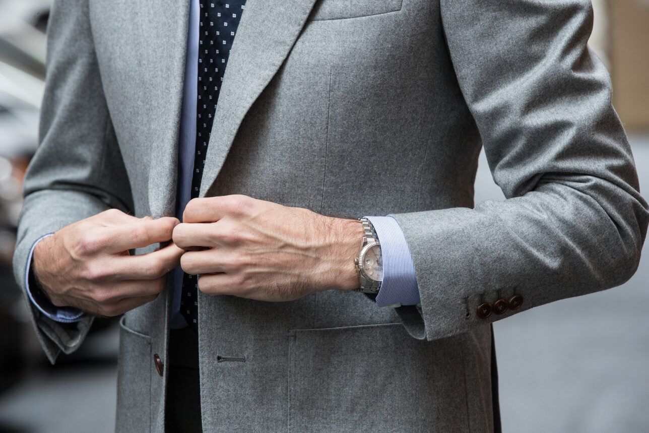 Grey flannel sport coat with peak lapel, light blue striped shirt and navy dotted tie, detail of a model buttoning it