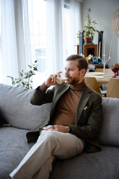 Olive green wool sport coat with brown turtleneck, cream trousers and pocket square, worn by a model seated on a sofa