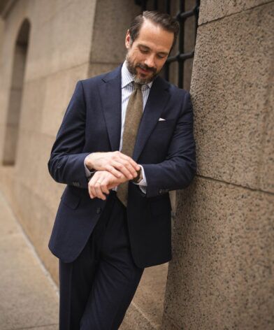 Navy flannel suit with blue striped shirt, brown wool tie and white pocket square, worn by a model leaning on a stone wall