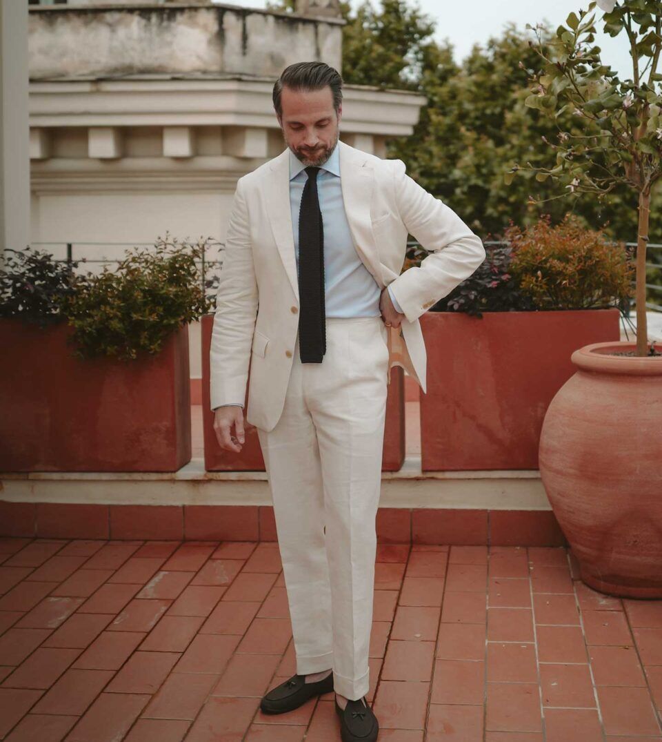 Cream linen suit with light blue shirt, navy knit tie and dark tassel loafers, worn by a model on a terrace