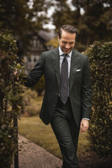 Forest green flannel suit with striped shirt, navy medallion tie and white pocket square, worn by a model in a garden