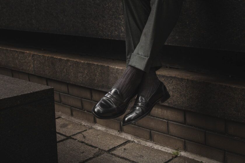Black leather penny loafers with ribbed dark socks and cuffed green flannel trousers, worn by a seated model