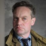 Navy silk necktie with gold motifs, blue chambray shirt and olive field jacket, worn by a man in a portrait
