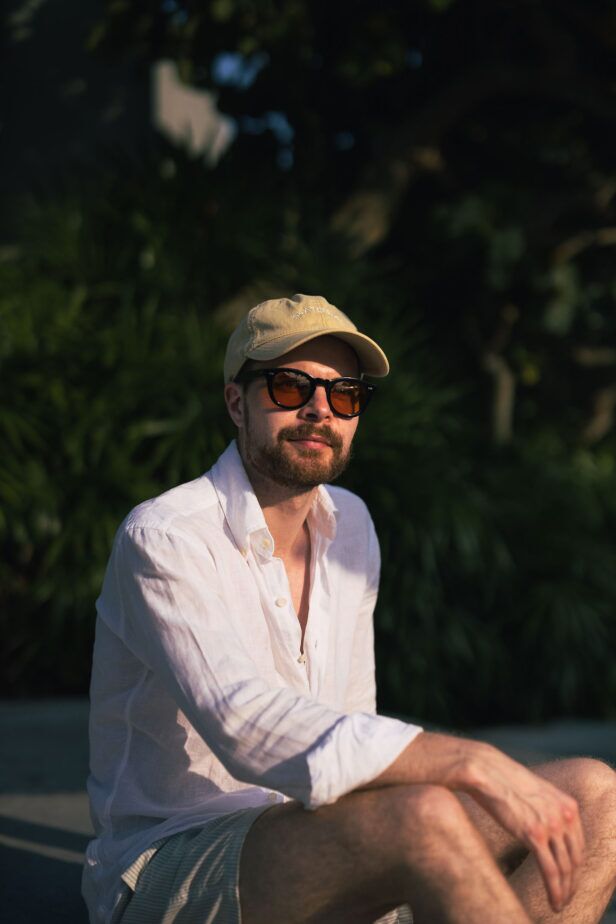 Orange-tinted sunglasses with black frames, worn with a white linen shirt and tan cap in a garden