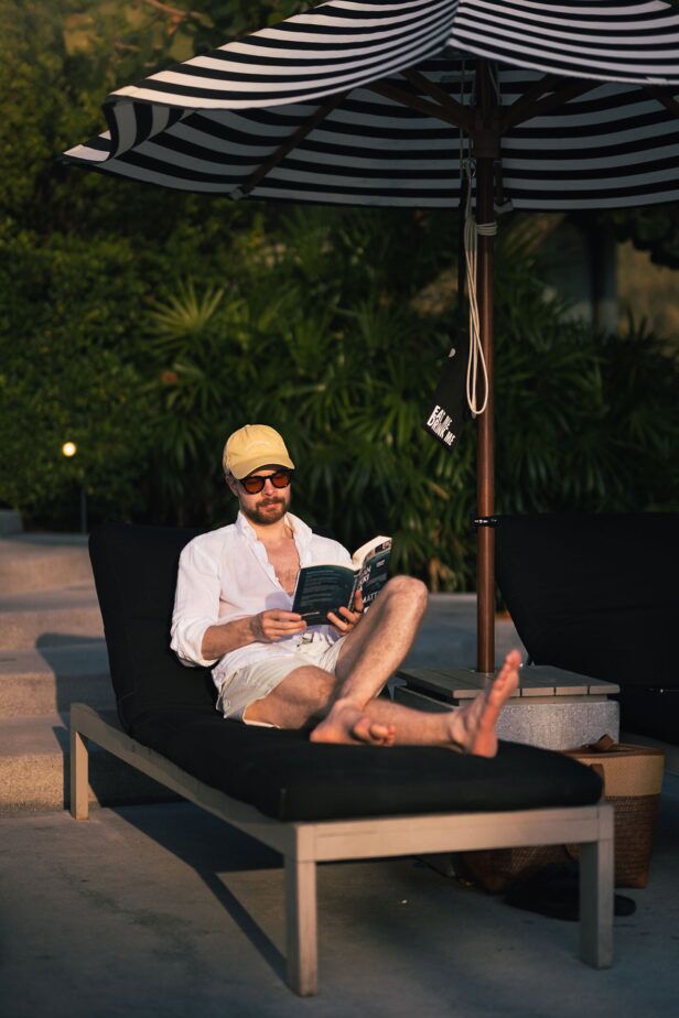 Tinted round sunglasses with white linen shirt, cream shorts and yellow cap, worn by a model reading on a poolside lounger