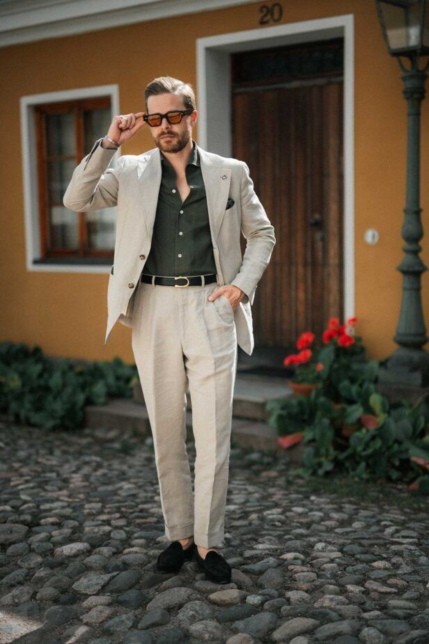 Cream linen suit with dark green shirt, black belt and black suede loafers, worn by a model on a cobbled street