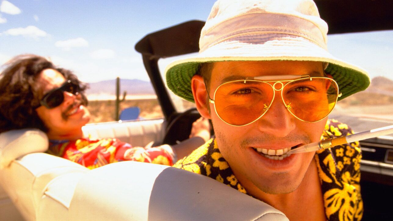 Yellow-tinted aviator sunglasses with white bucket hat and yellow floral shirt, worn in a convertible, film still