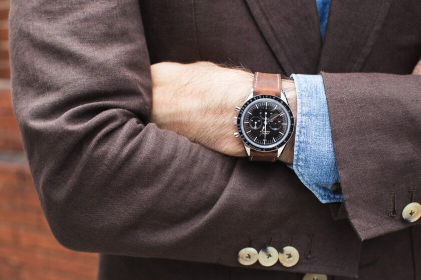 Black-dial chronograph watch on tan leather strap with brown linen sport coat and denim shirt cuff, a close-up detail
