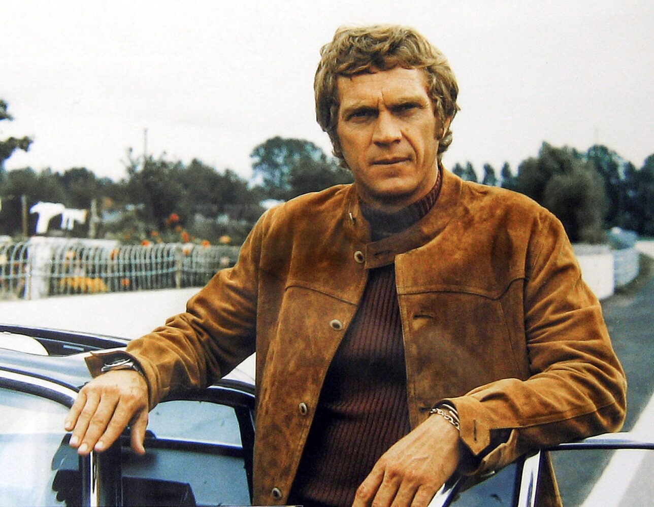 Tan suede blouson jacket with band collar over a burgundy ribbed turtleneck, worn leaning on a car in a lifestyle scene