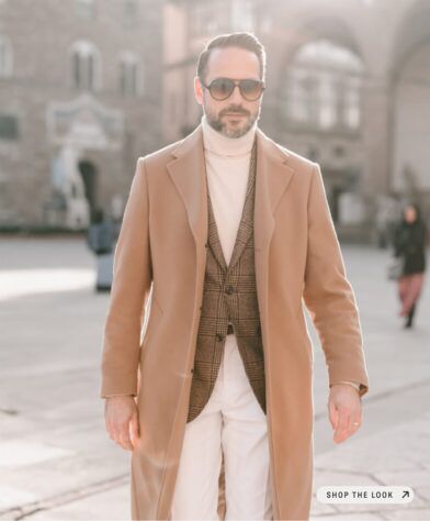 Camel wool overcoat over a brown glen check sport coat, cream turtleneck and trousers, worn by a model on a sunlit piazza
