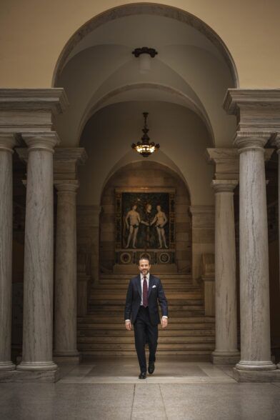 Navy chalk stripe suit with white shirt, burgundy dot tie and white pocket square, worn by a model in a marble hall