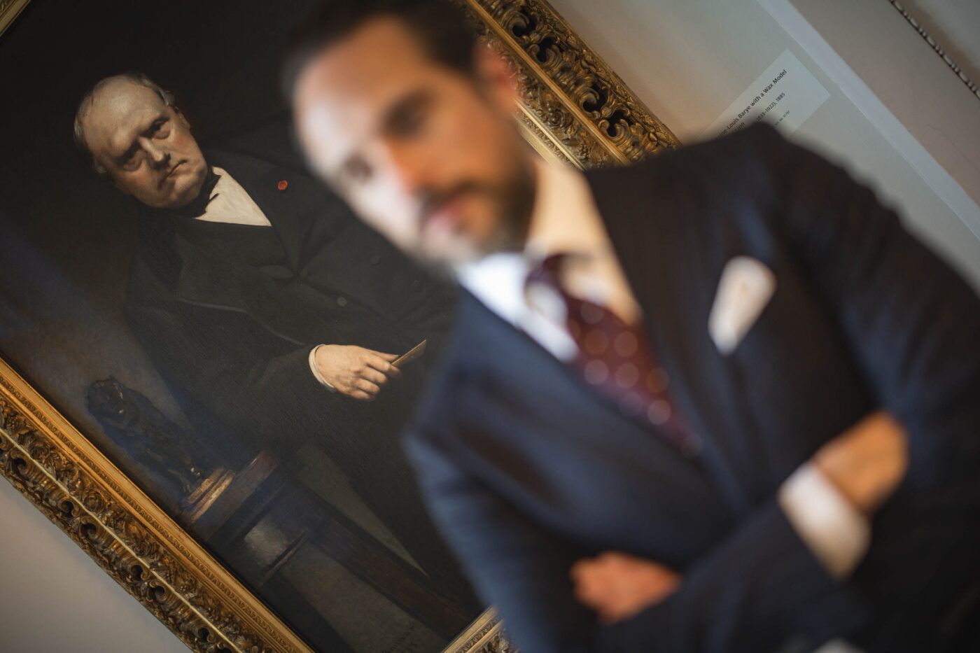 Navy suit with burgundy dot tie and white pocket square, worn by a model in a gallery beside a framed portrait