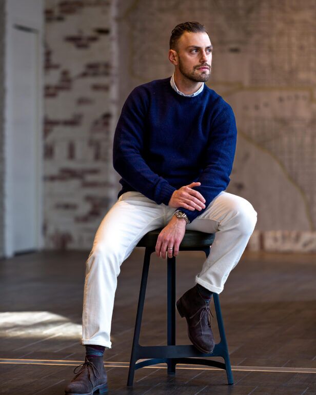 White denim jeans with navy crewneck sweater and brown suede chukka boots, worn by a seated model