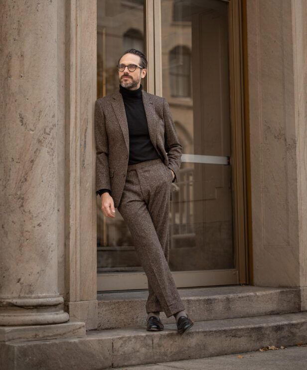 Brown flecked donegal tweed suit with black turtleneck and black loafers, worn by a model on stone steps