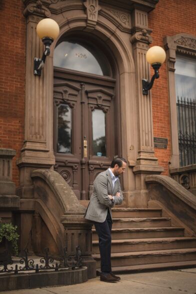 Grey tweed sport coat with white shirt, navy trousers and brown suede loafers, worn by a model on brownstone steps