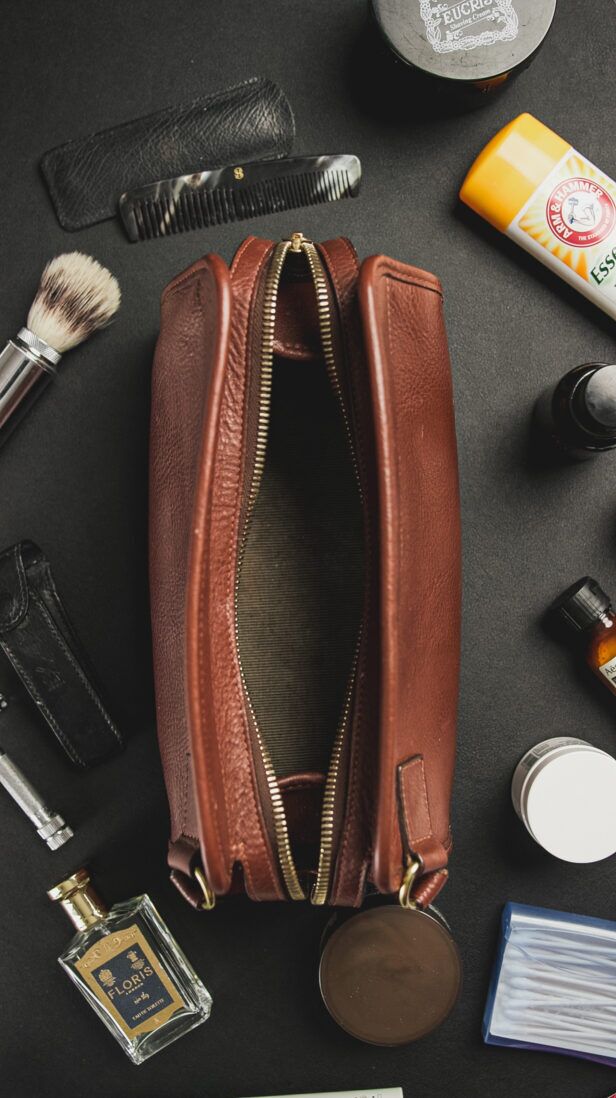 Brown leather zip dopp kit shown open, surrounded by shaving brush, comb and grooming bottles in a styled flat lay