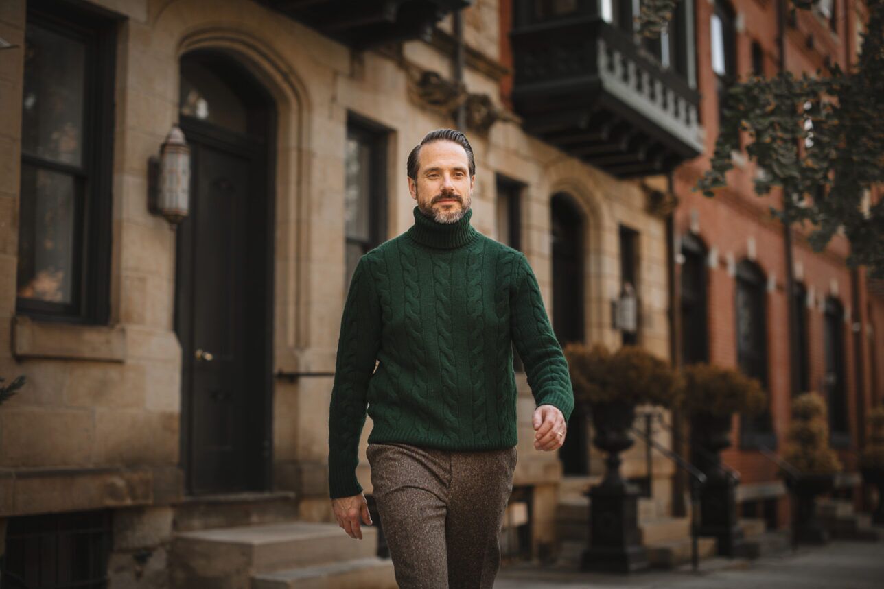 Green cable knit wool turtleneck sweater with brown tweed trousers, worn by a model on a city street