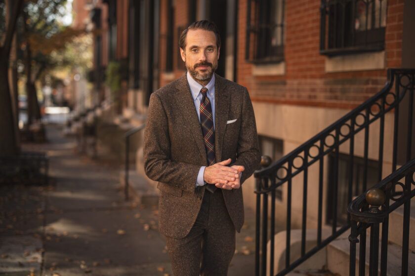 Brown donegal tweed suit with light blue shirt, plaid tie and white pocket square, worn by a model on a city street